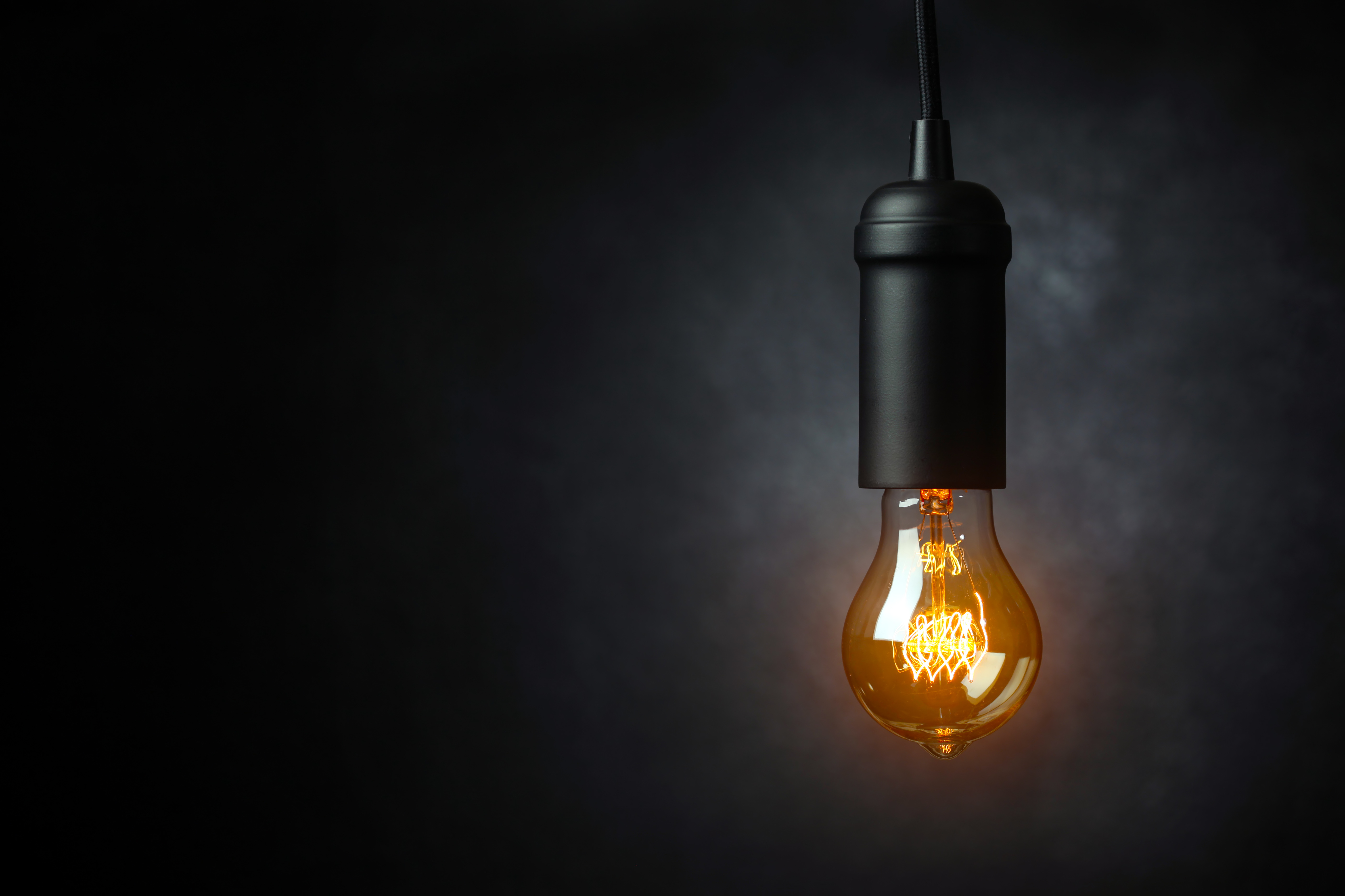 A bare incandescent lightbulb shines in front of a dark gray background.