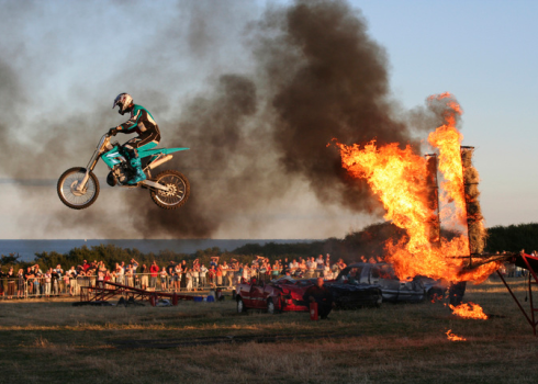 A crowd looks on as a motorcycle stunt driver soars through the air having jumped through a ring of fire.